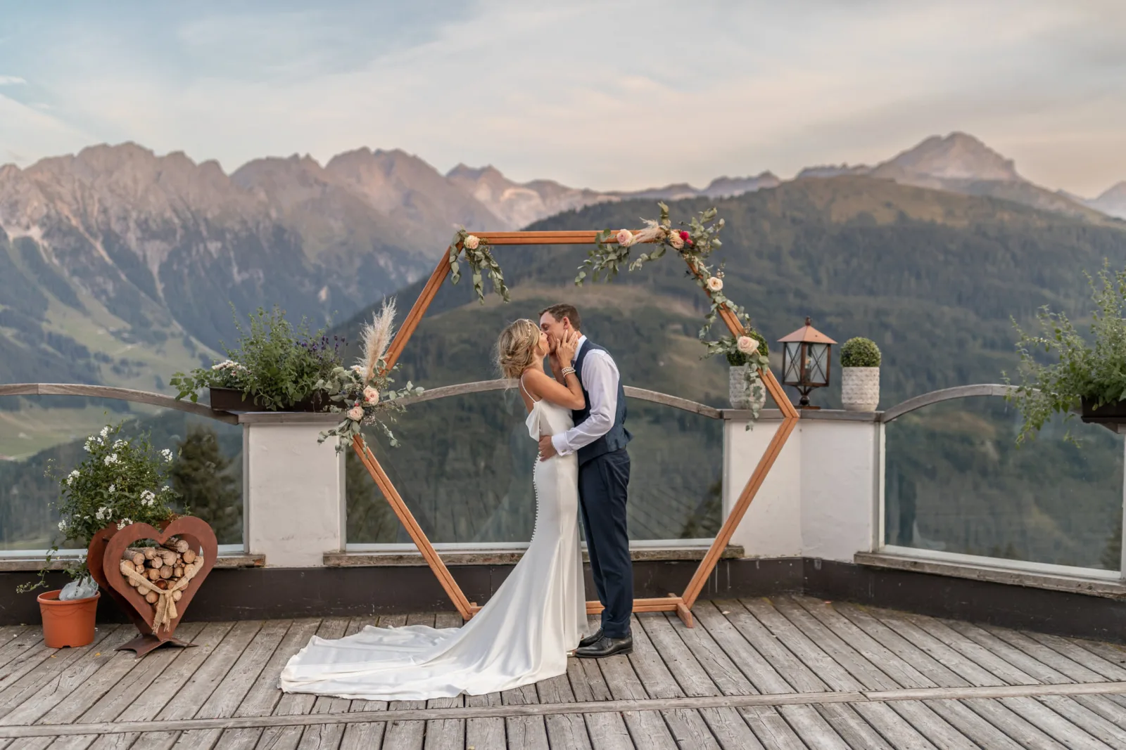 Bride and groom kissing beneath a wooden hexagonal arch on a mountain terrace in the Austrian Alps, with peaks behind