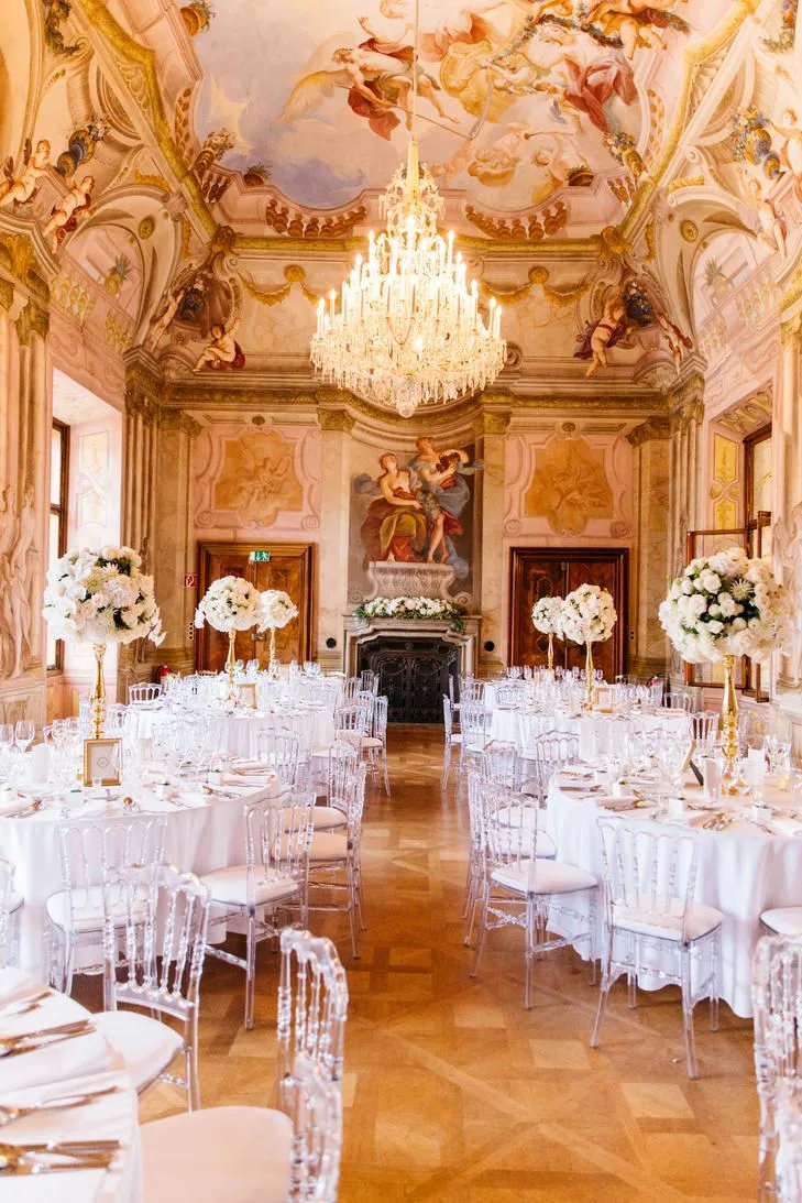 Wedding reception in a baroque palace hall in Austria, with painted ceiling frescoes, a crystal chandelier and round tables with tall white flower arrangements