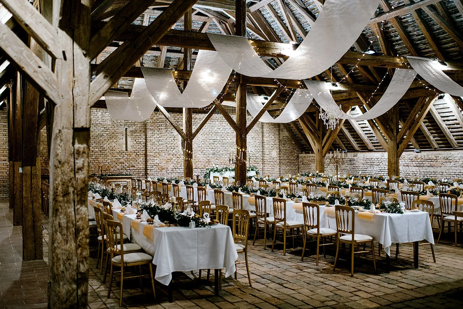 Rustic barn wedding reception in Austria with exposed timber beams, brick walls, draped fabric, fairy lights and long white tables