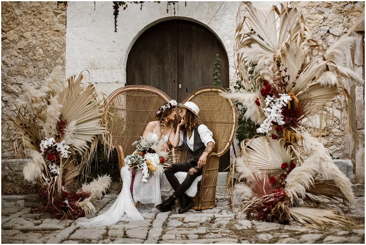 Boho wedding couple kissing on peacock wicker chairs framed by dried palm leaves, pampas grass and red roses in a stone courtyard