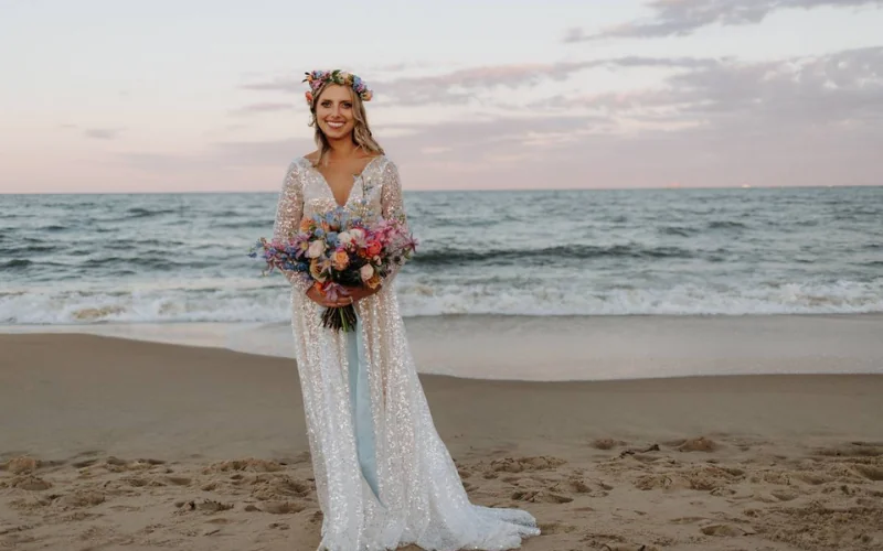A bride on the beach at dusk wearing a fresh flower crown and holding a brightly coloured bouquet