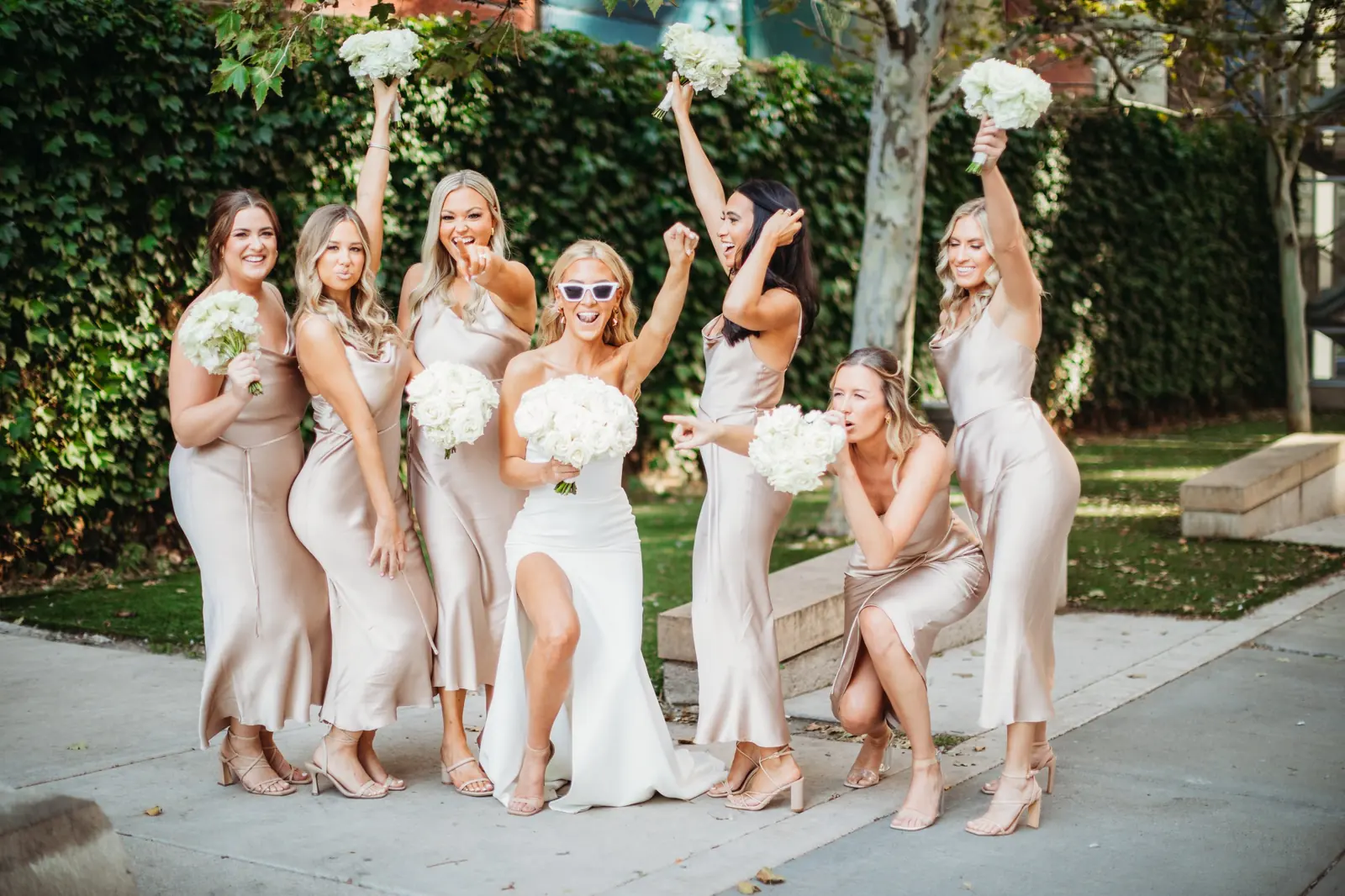 Bride in sunglasses celebrating with bridesmaids in champagne satin dresses, holding white rose bouquets in the air
