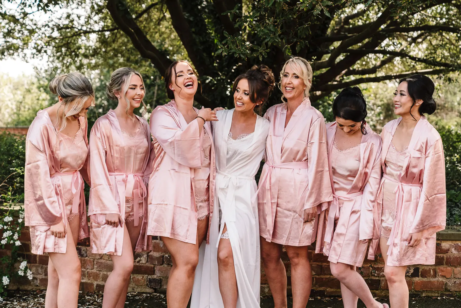 Bride in a white robe laughing with her bridesmaids in pink satin robes on the wedding morning, a getting-ready moment for a content creator
