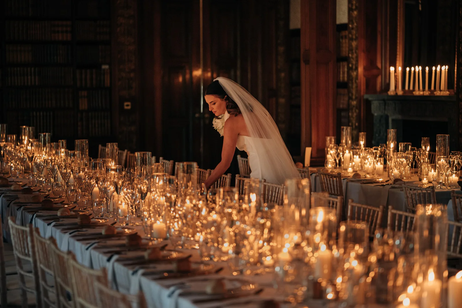 Bride adjusting a candlelit wedding reception table in a wood-panelled library, with hundreds of candles and glasses glowing