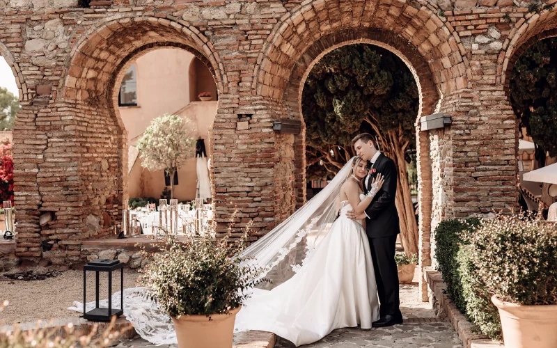 A bride and groom embracing beneath brick archways at a Spanish wedding venue, her long veil trailing across the courtyard