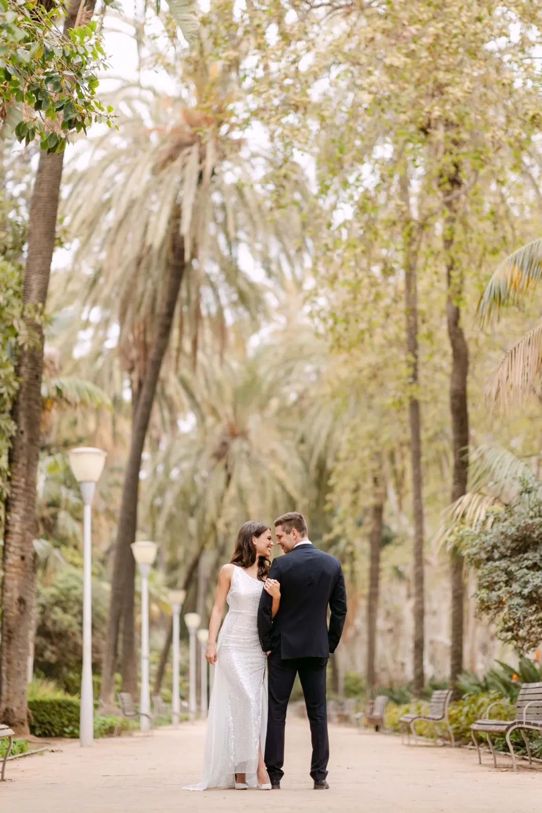 Bride in a sequinned gown and groom sharing a smile on a palm-lined promenade, an editorial-style wedding photograph