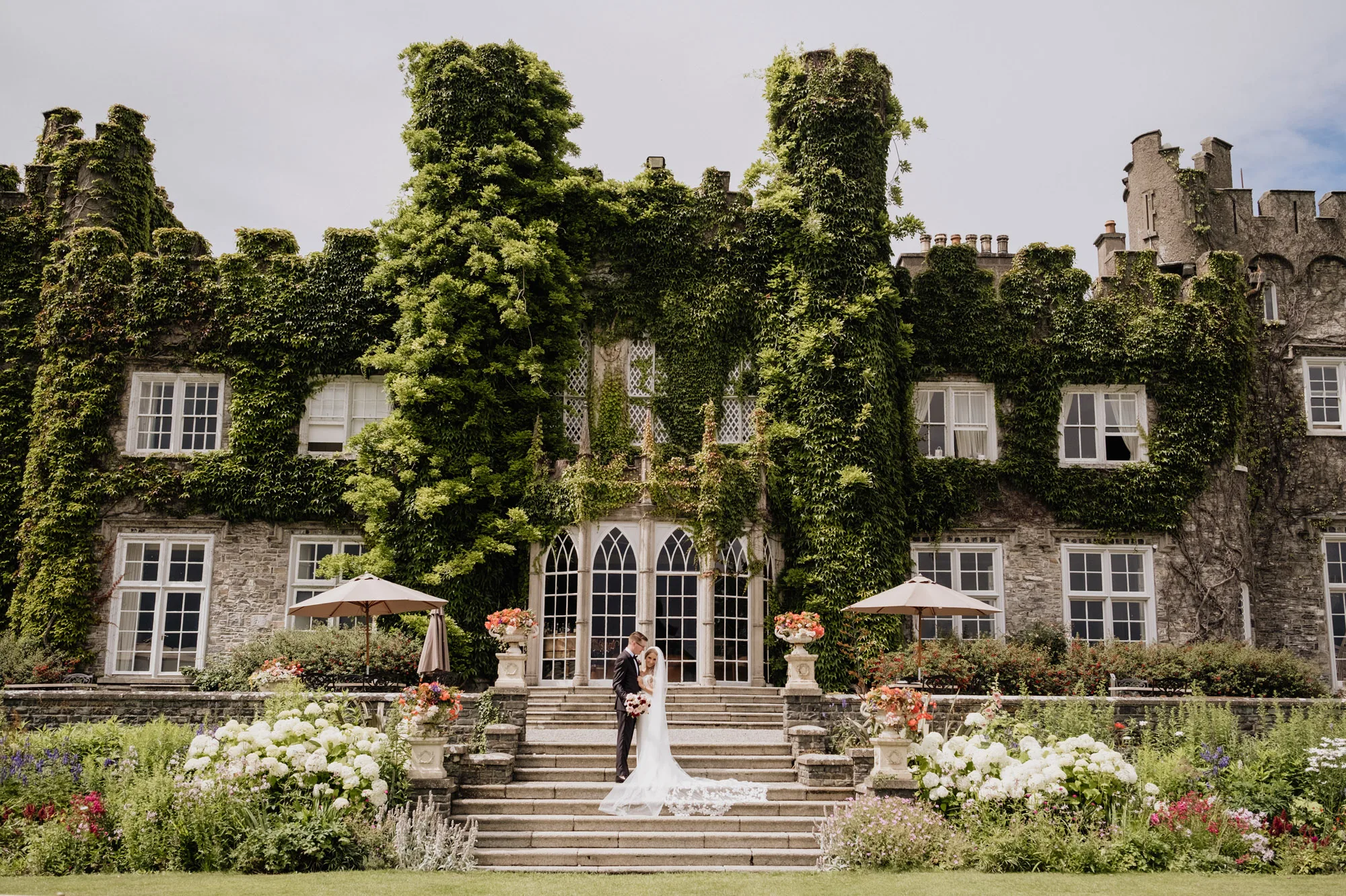 Bride and groom on the garden steps of an ivy-covered castle wedding venue in Ireland, framed by white hydrangeas