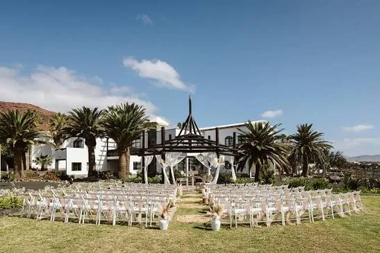 Outdoor wedding ceremony with white chairs and a draped pergola on a lawn, with palm trees and a whitewashed building in the Canary Islands