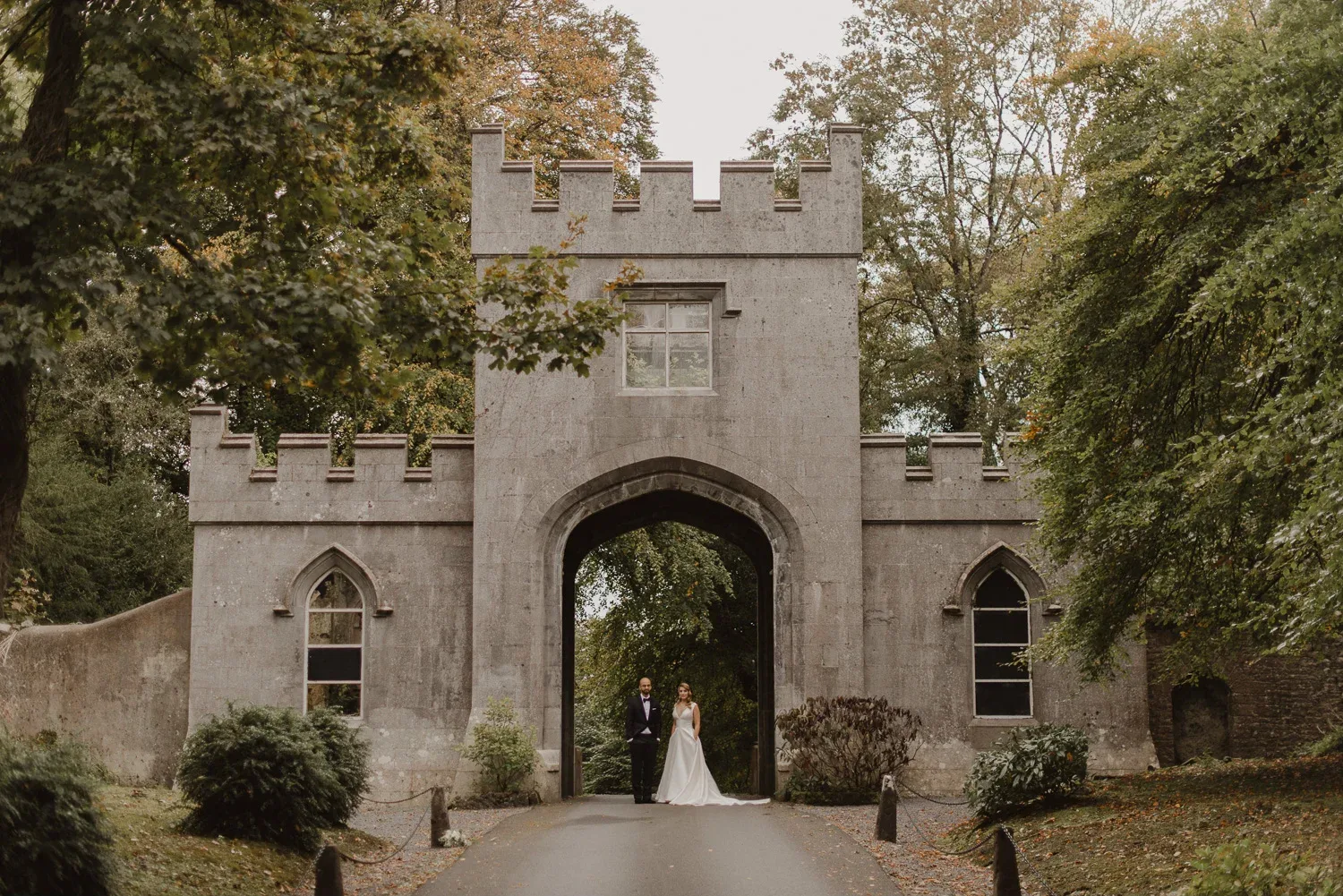 Bride and groom standing beneath the arch of a crenellated castle gatehouse in Irish woodland