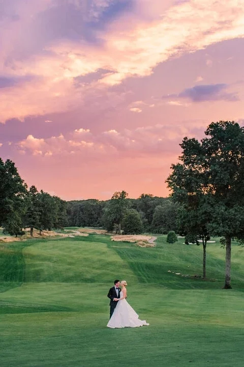 Bride and groom embracing on a golf course fairway under a pink and gold sunset sky