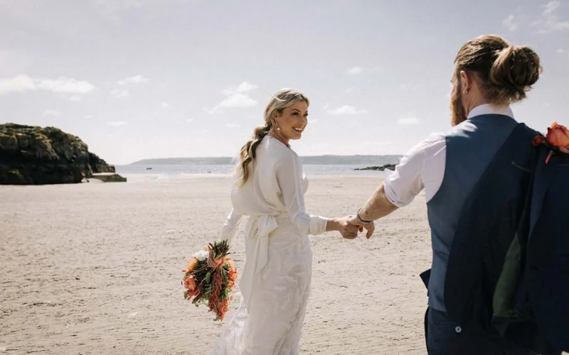 A bride leading her groom across the sand, her hair worn in a low secured ponytail, holding a coral bouquet