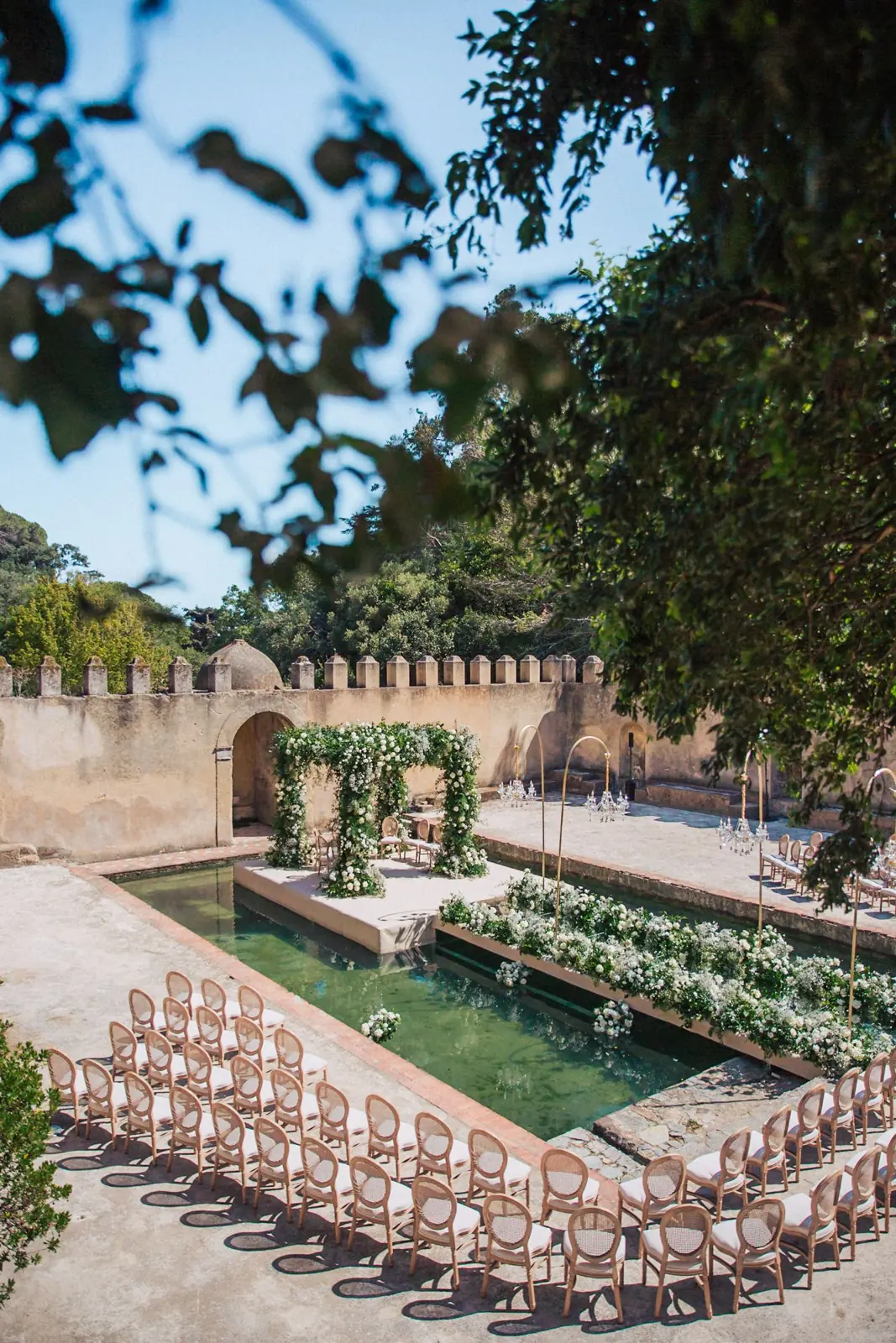 Wedding ceremony set beside a reflecting pool in a walled courtyard, with a white floral arch, chandeliers and curved rows of rattan chairs