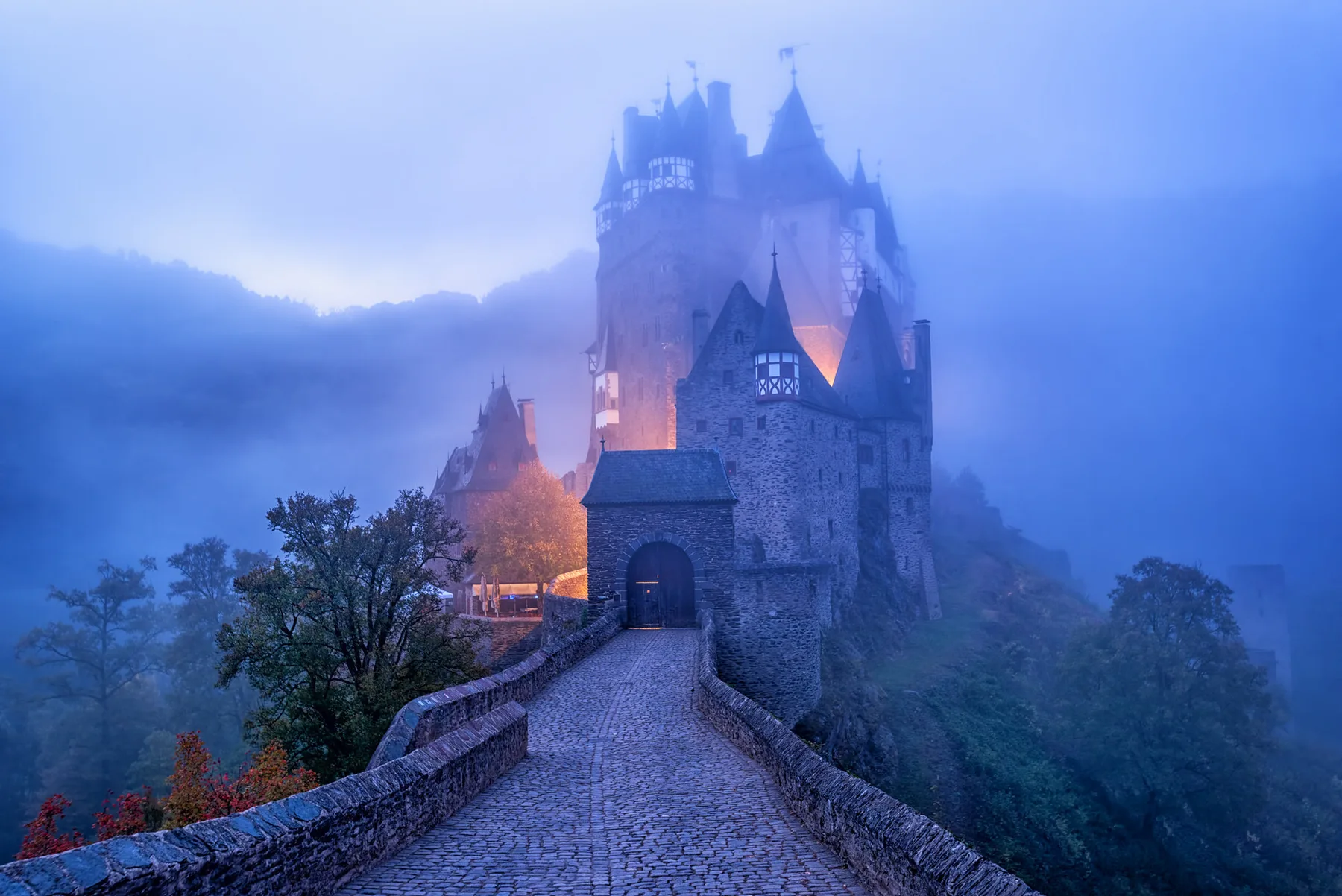 Medieval German castle with turrets and half-timbered towers rising out of the morning mist in the Moselle region, with a cobbled bridge leading to the gatehouse