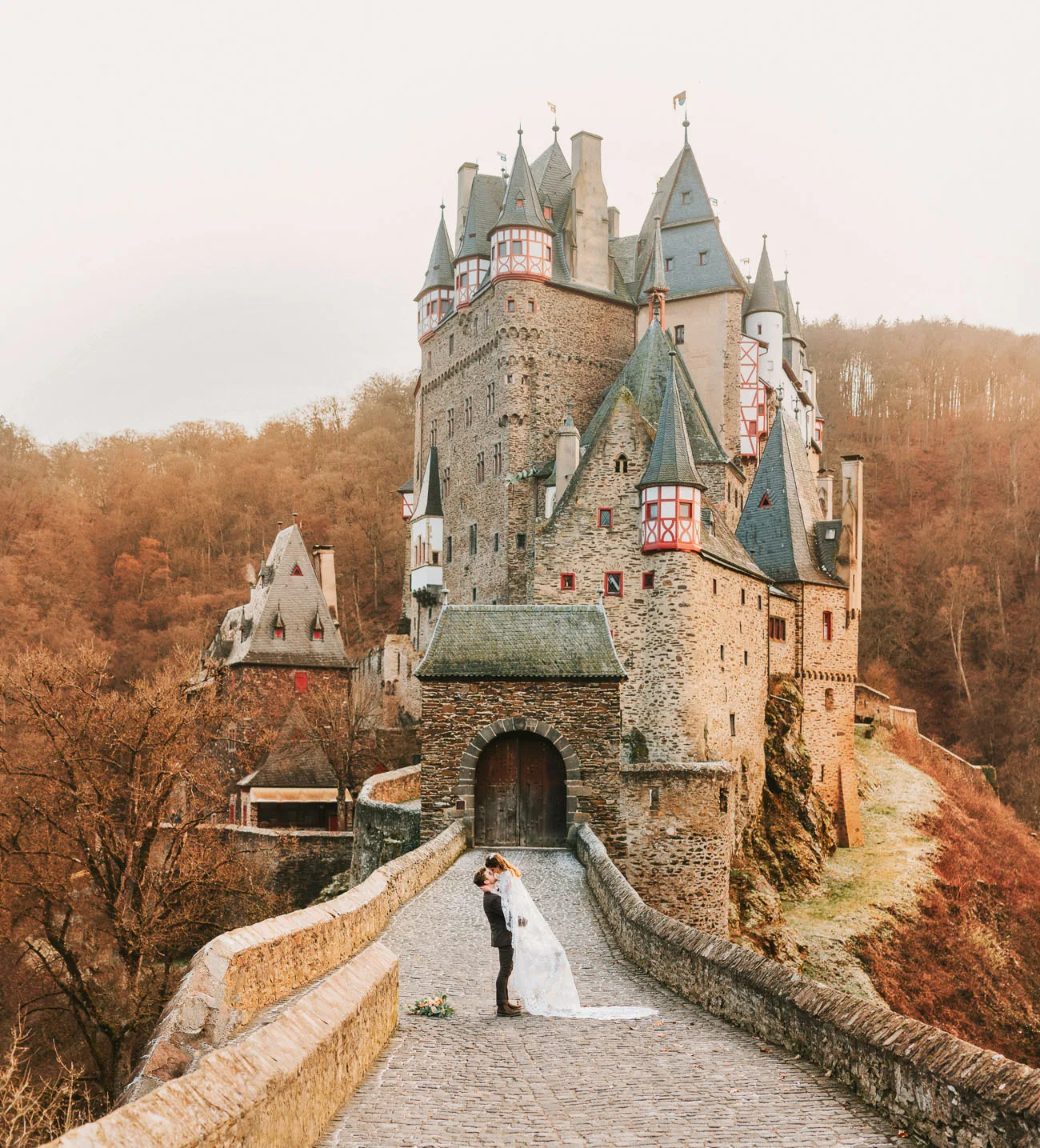 Bride and groom kissing on the cobbled bridge of a medieval castle in the Moselle region of Germany, surrounded by autumn woodland