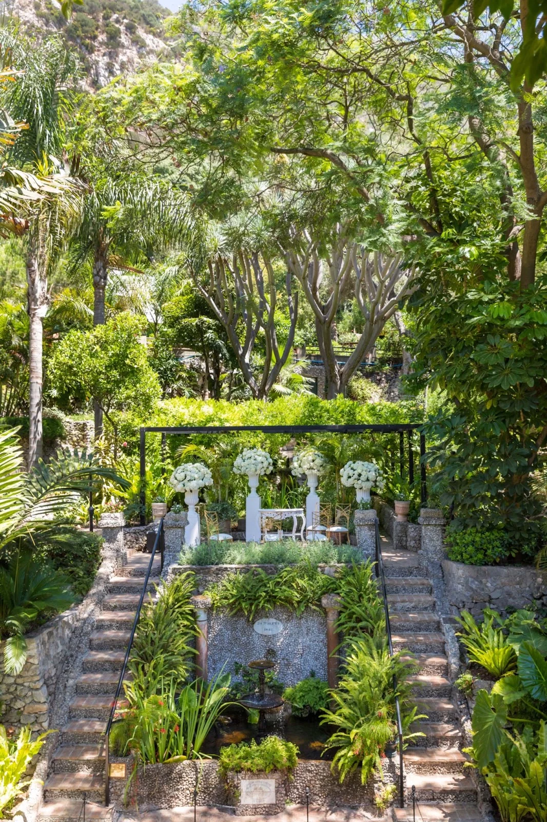 Wedding ceremony set up beneath a pergola with white flowers in botanical gardens in Gibraltar, with stone steps, palms and the Rock behind