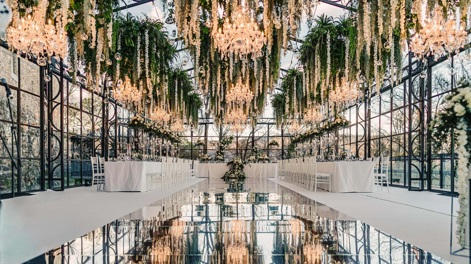 Glasshouse wedding reception with crystal chandeliers, cascading white hanging flowers, long white tables and a mirrored aisle