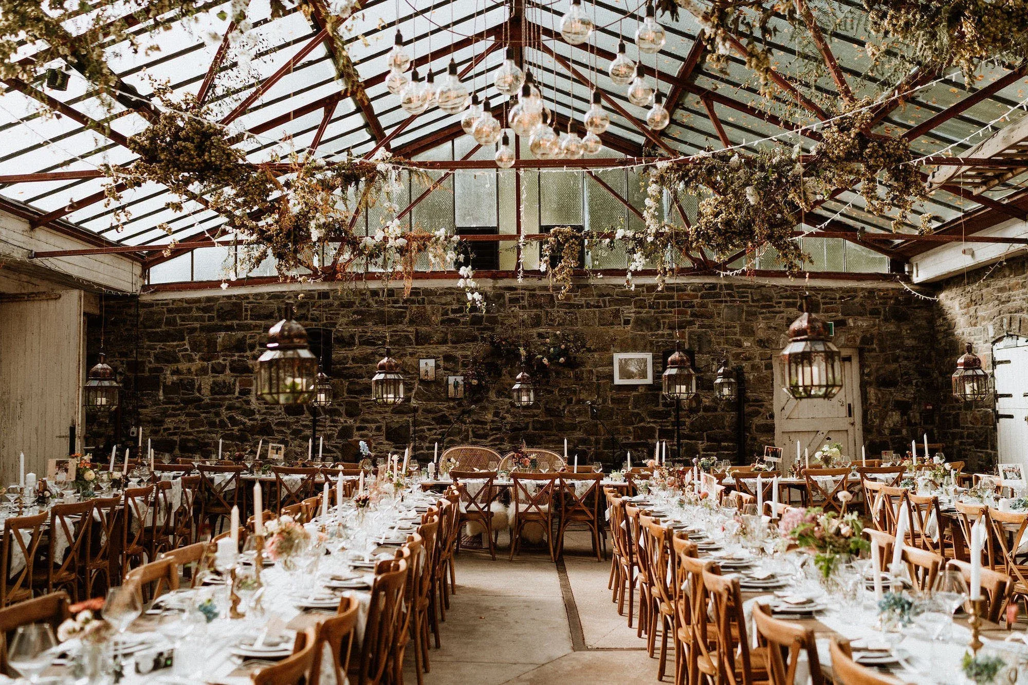 Wedding reception in a Welsh glasshouse venue with stone walls, hanging dried flowers, lanterns and long candlelit tables