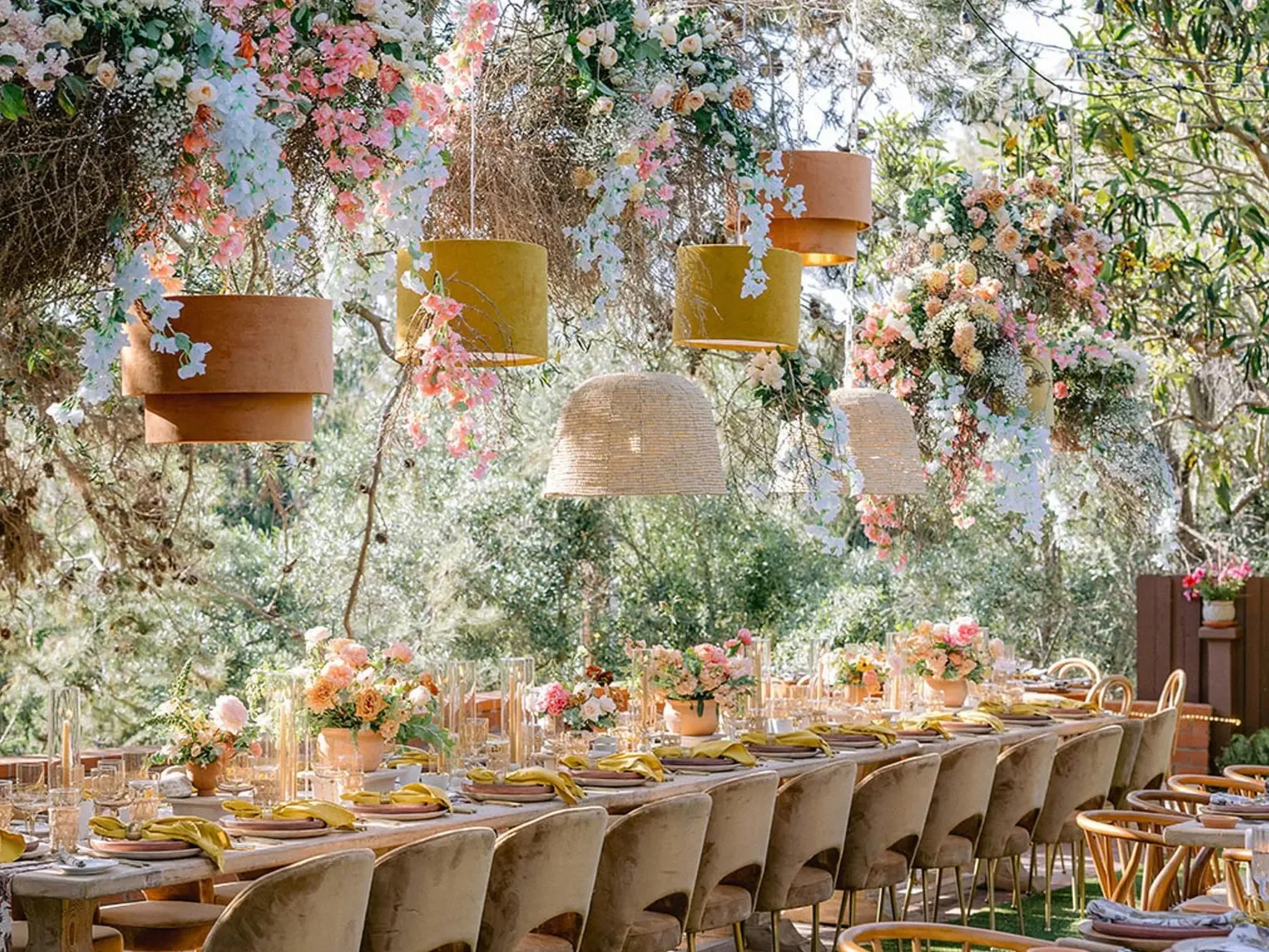 Velvet and rattan lampshades suspended among hanging floral installations above a long wedding table