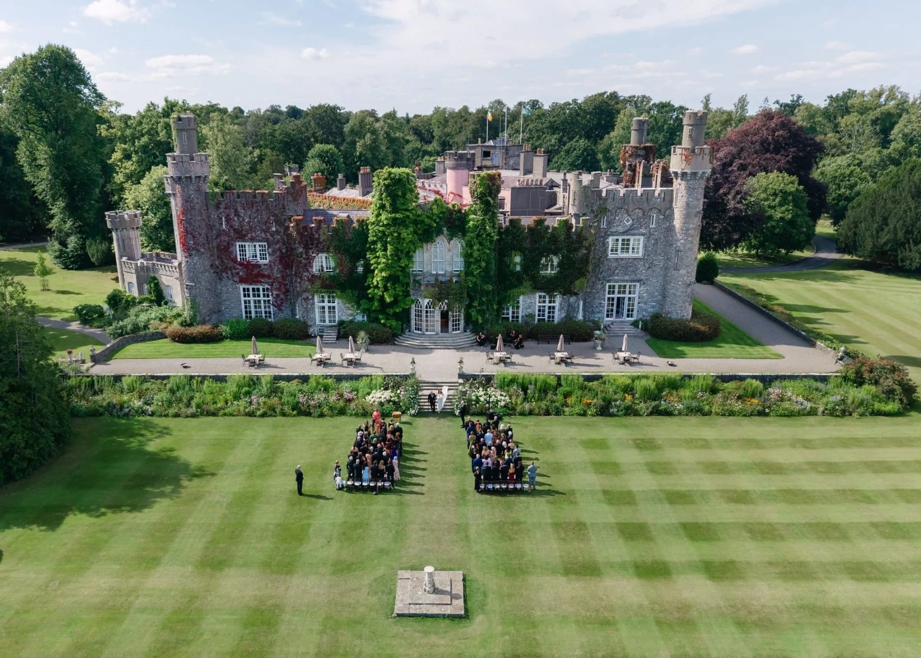 Aerial view of an outdoor wedding ceremony on the striped lawn of a turreted Irish castle wedding venue