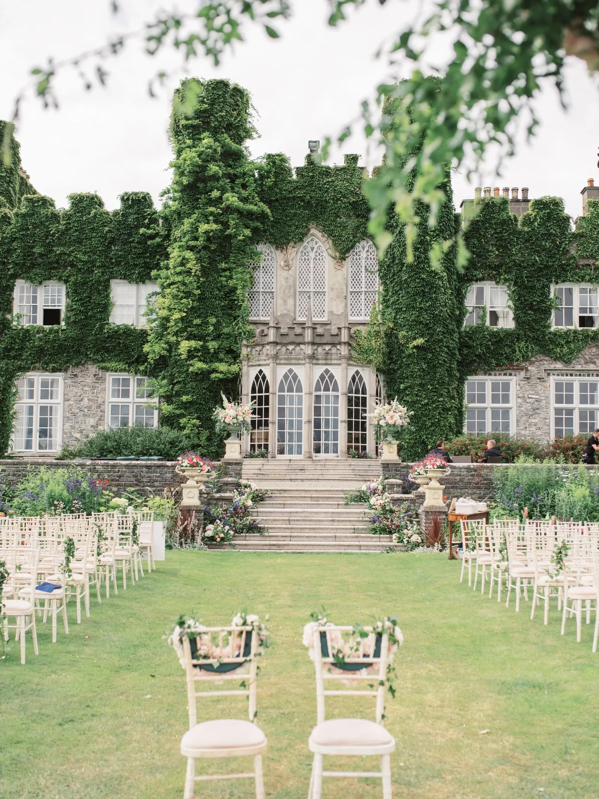 Outdoor wedding ceremony set up on the lawn of an ivy-covered Irish castle, with floral urns and white chairs
