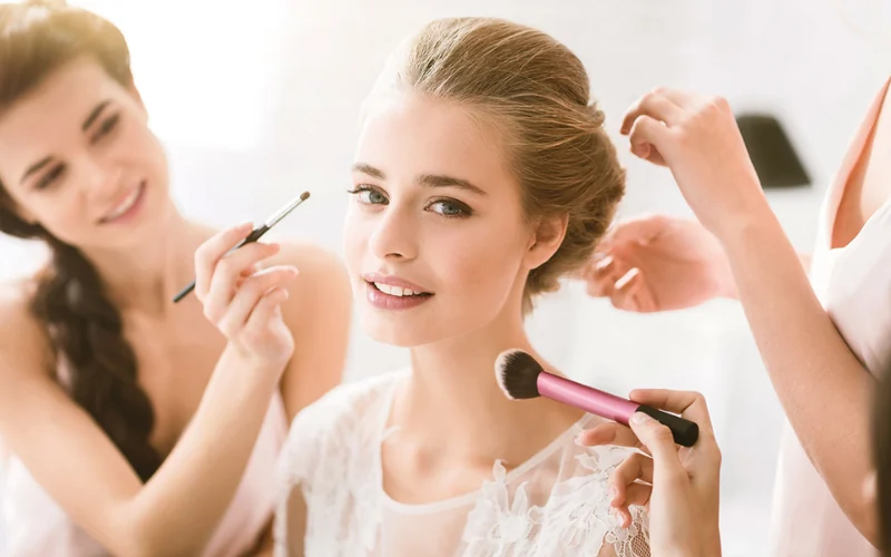 A wedding makeup artist applying eye makeup while a second stylist works on the bride's updo during the morning preparations
