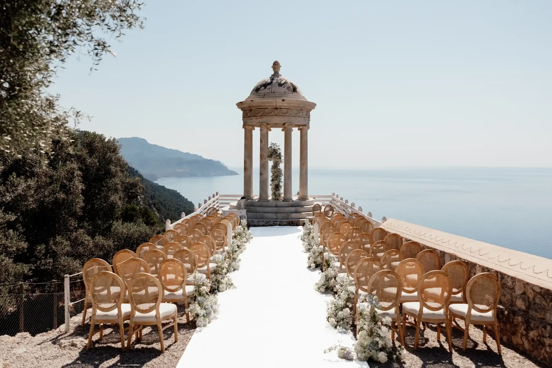 Clifftop wedding ceremony in Mallorca with a domed stone temple, rattan chairs and a white aisle above the Mediterranean