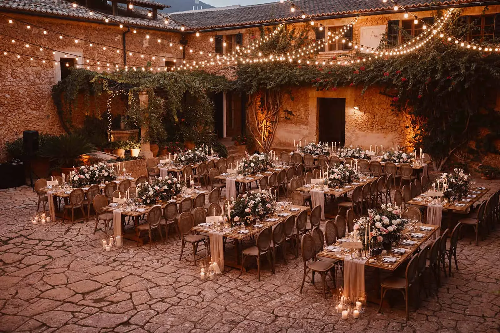 Long candlelit wedding dinner tables under festoon lights in the stone courtyard of a Mallorcan finca