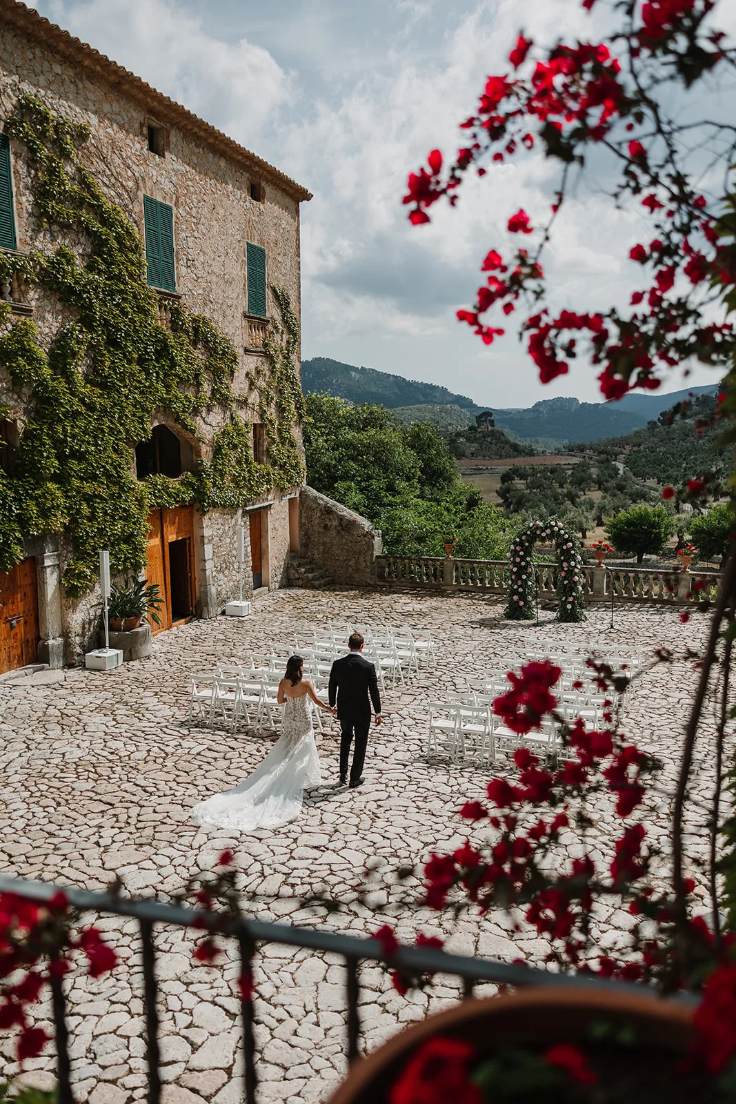Bride and groom walking to their ceremony in the cobbled courtyard of an ivy-covered stone finca in the Mallorcan mountains