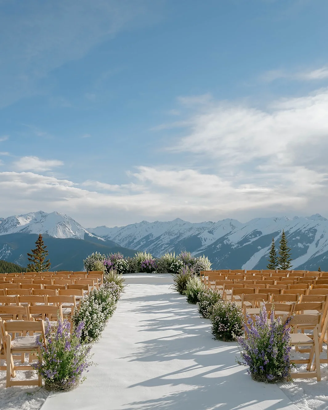 Outdoor wedding ceremony aisle lined with wildflowers and wooden chairs, with snow-capped mountain peaks behind