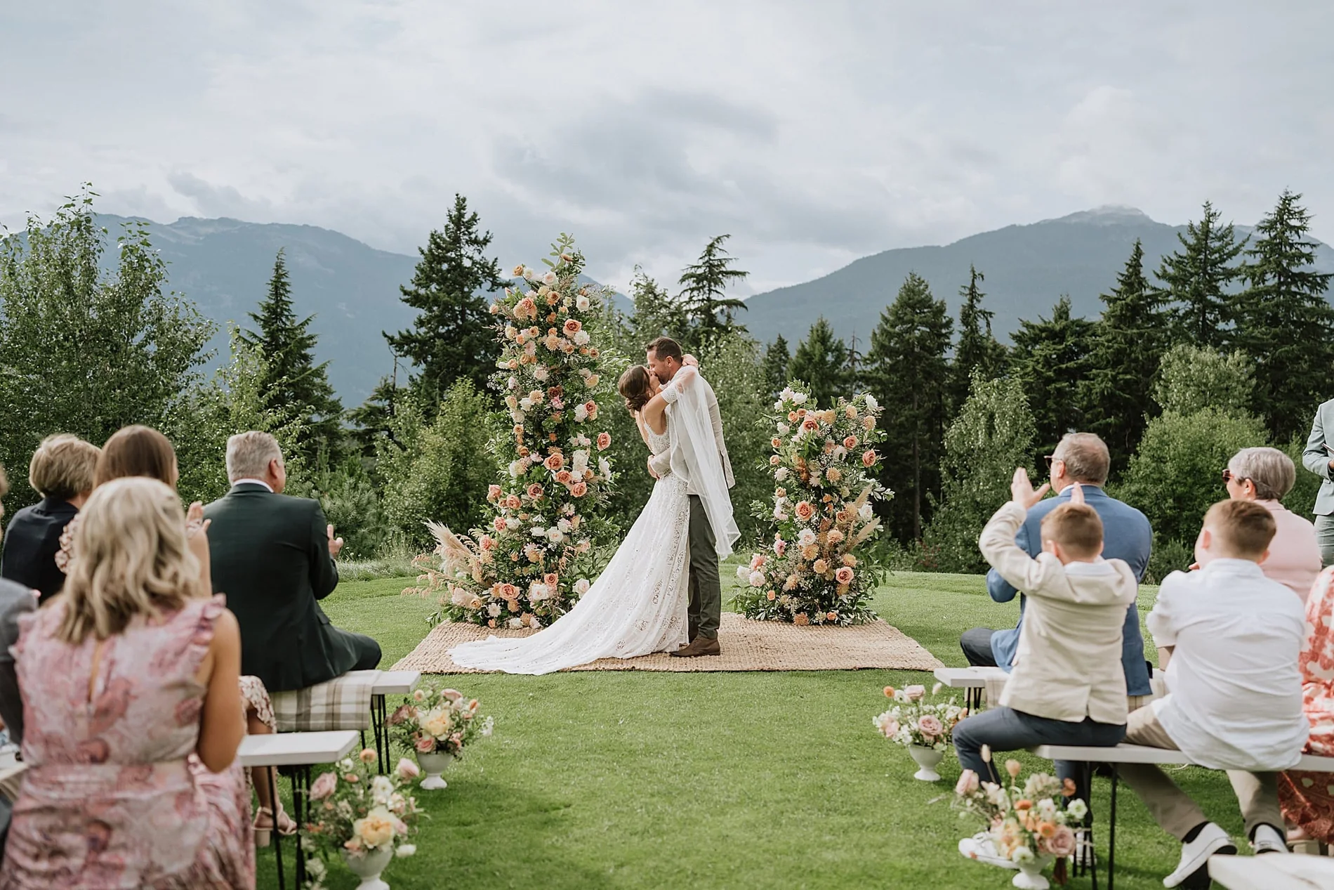 Bride and groom sharing their first kiss between tall floral pillars of white flowers and greenery at an outdoor wedding ceremony