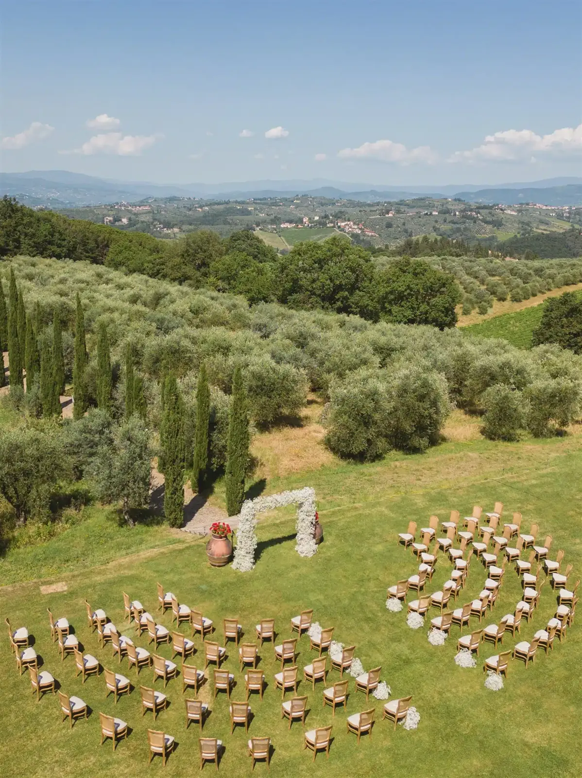 Aerial view of an outdoor hillside wedding ceremony with a floral arch, surrounded by trees and rolling hills