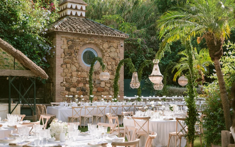 Round tables set for an outdoor wedding reception in front of a stone finca in Spain, with chandeliers and greenery hung from the palms