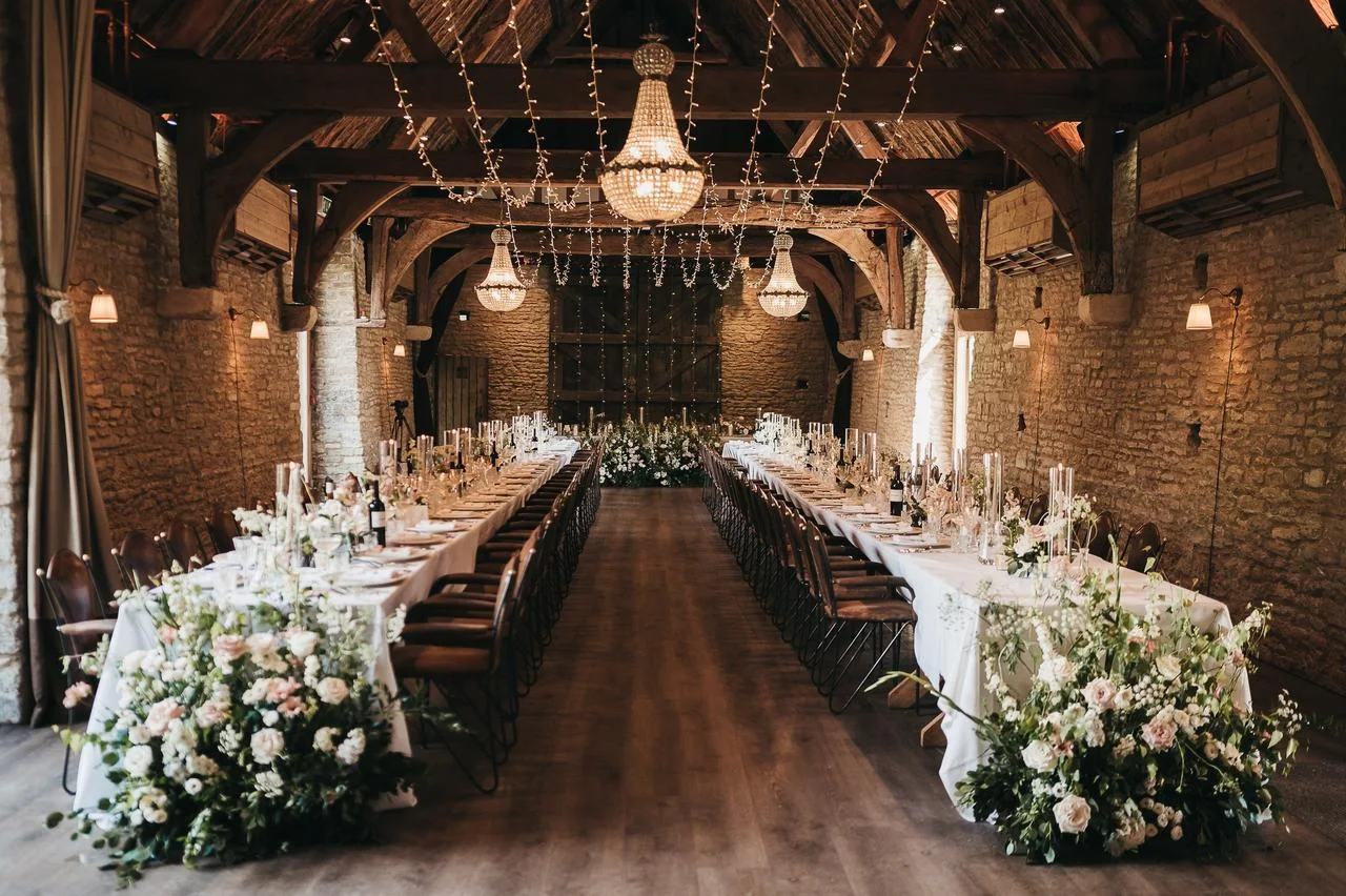 Long winter wedding reception tables in a stone barn with exposed beams, crystal chandeliers, fairy lights and white flowers