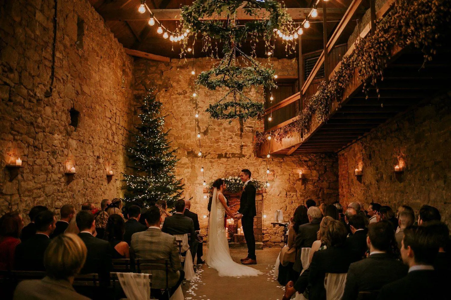 Winter wedding ceremony in a stone barn lit with fairy lights, candles, hanging greenery and a Christmas tree