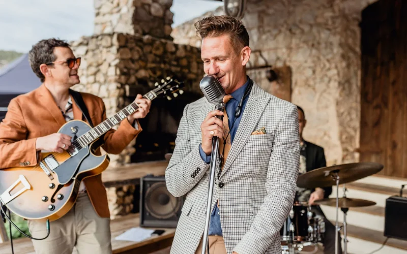 A wedding band's singer and guitarist performing beside a stone fireplace at an outdoor drinks reception