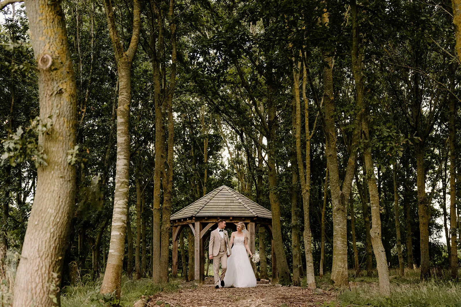 Bride and groom walking from a wooden gazebo through woodland at an English wedding venue