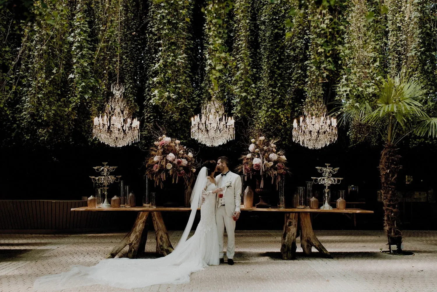 A bride and groom beside a floral table lit by crystal chandeliers, an on-the-day wedding coordination by Swish Soirées