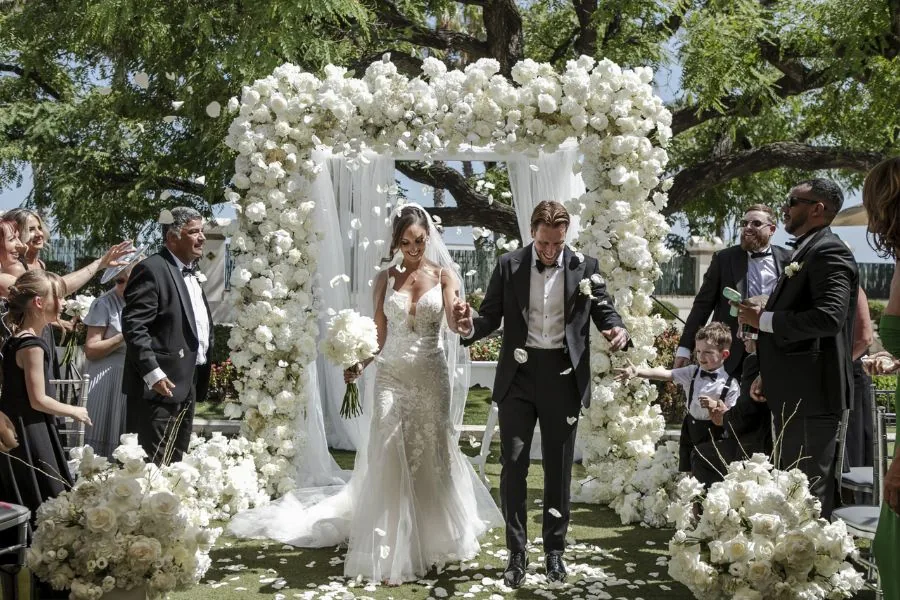 Newlyweds walking through a white floral arch amid confetti, a luxury wedding planned by Swish Soirées